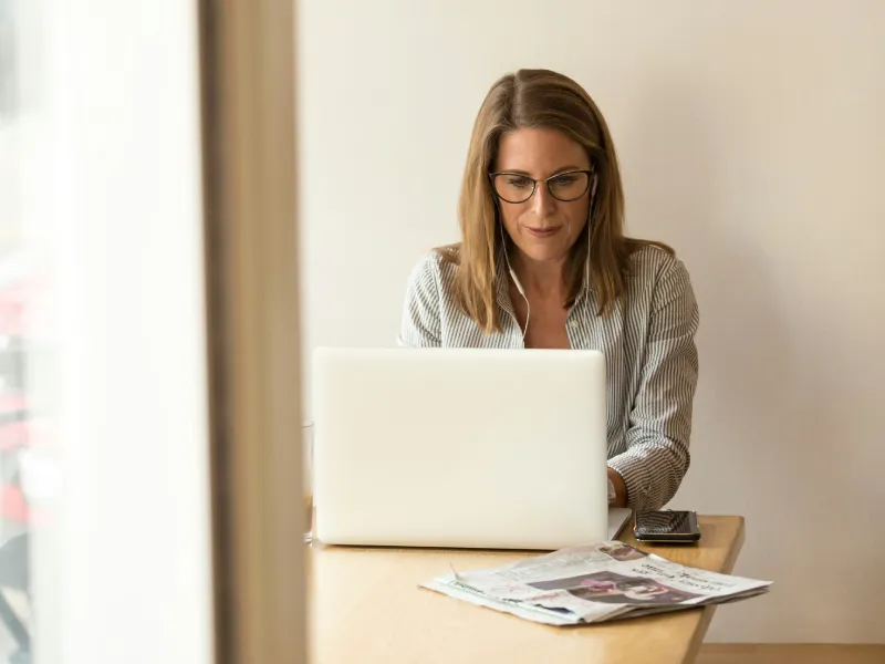 Focused woman wearing glasses works on a laptop at a wooden table, with newspapers and a smartphone nearby in a bright, calm room.